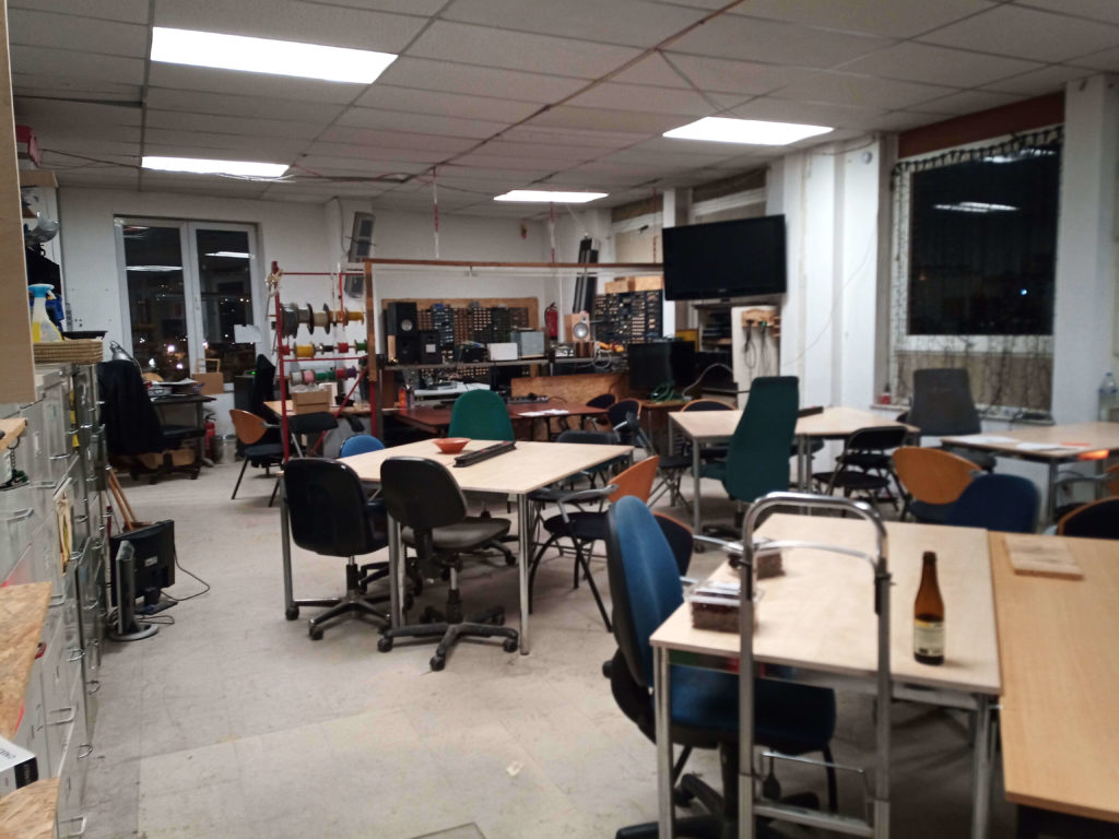 Photo of Hackerspace Brussels' main room featuring tables, chairs and an electronics workspace at the far end.